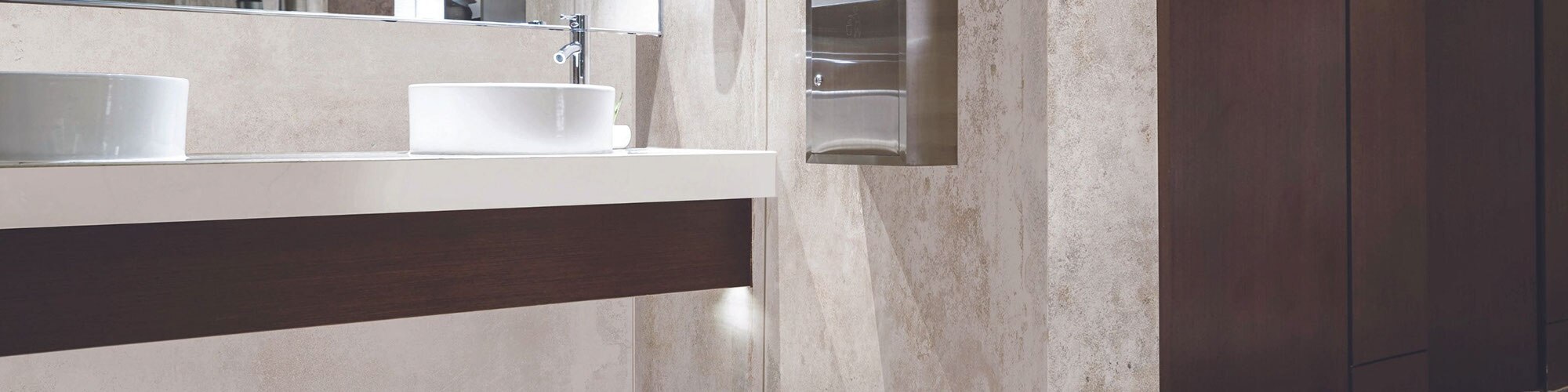 Office building bathroom with off-white & beige porcelain slab wall and backsplash, floating vanity of wood with white quartz countertop and vessel sinks, and dark wood stall doors.