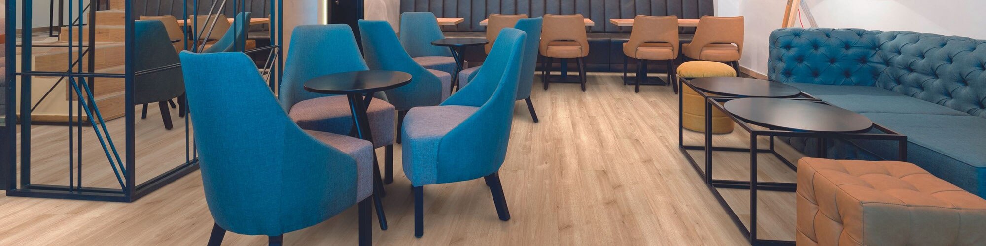 Restaurant dining room with wood look LVT (luxury vinyl tile) flooring, tables and blue cushioned chairs, blue sofa with coffee table and ottoman.