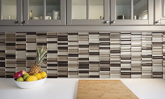 Varigated taupe, brown, and beige glass mosaic kitchen backsplash with gray cabinets and white countertop with a bowl of fruit.