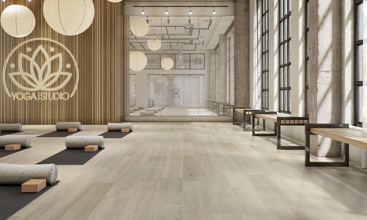 Yoga studio with LVT flooring that looks like wood planks, mirrored wall, sign with lotus flower, floor mats with pillows and blocks, and wooden benches against windows.