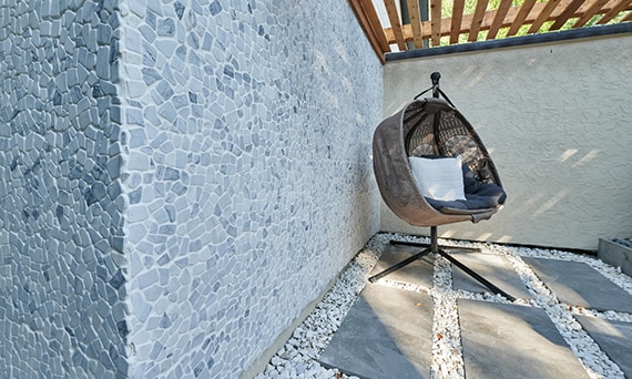 Outdoor patio with gray stone look 2CM porcelain pavers set in white gravel, wicker hanging chair on stand, and gray marble pebble mosaic wall tile.