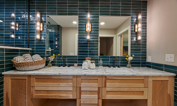 Bathroom with teal glazed porcelain wall tile, marble countertop with dual undermount sinks & mirrors, wood cabinets, and three glass & brass wall sconces.
