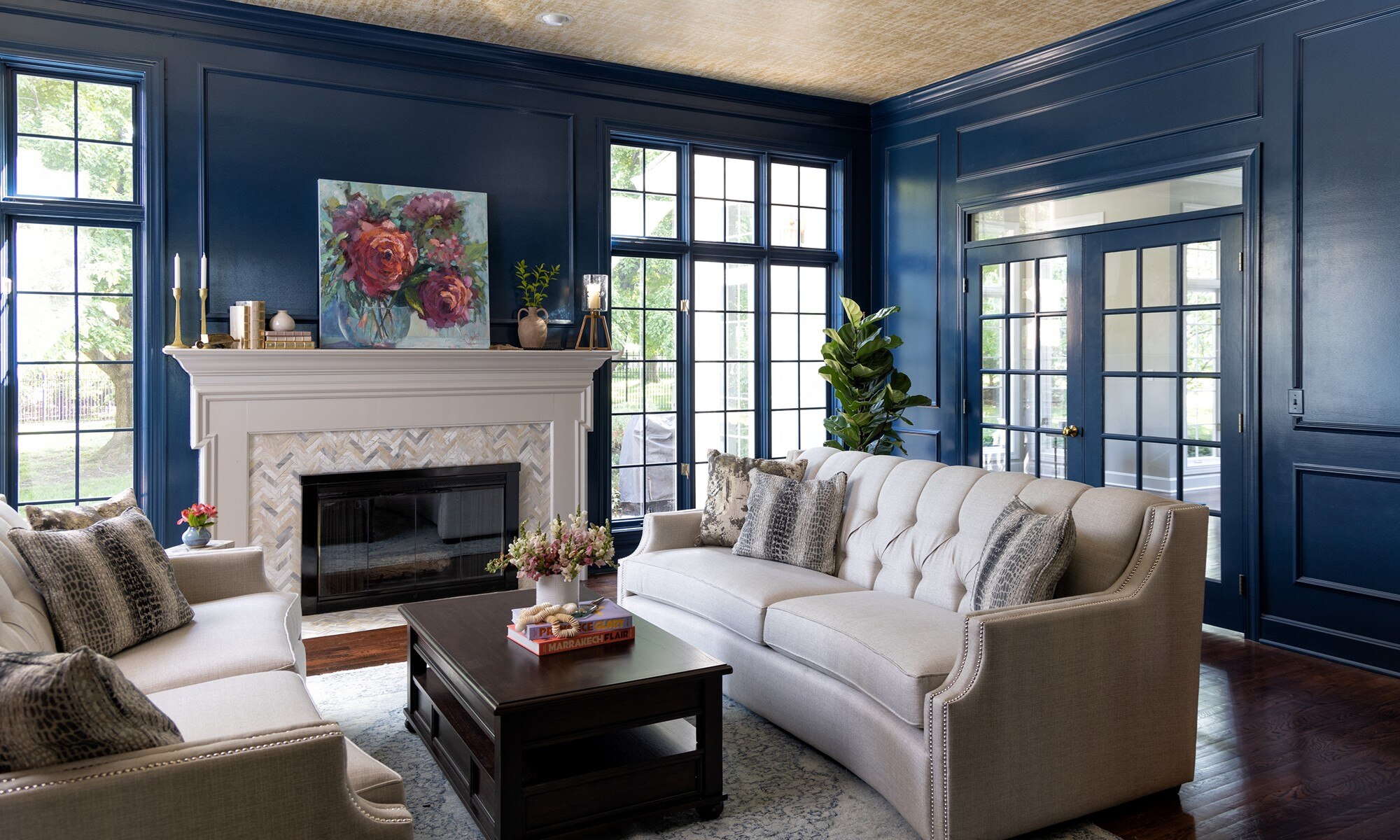 Renovated living room with dark blue paneled walls, floor-to-ceiling windows, fireplace with gray & beige marble mosaic surround & white mantel.