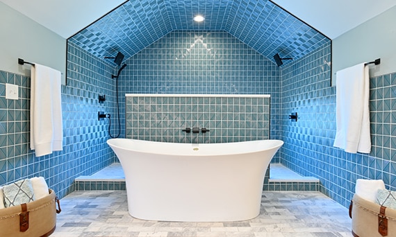 Renovated bathroom with soaker tub on gray marble floor tile, in front of large shower with textured blue geometric tile on the wall and ceiling.