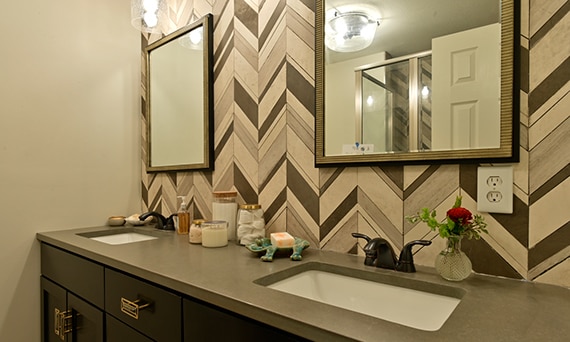 Bathroom vanity with brown quartz countertop, dual undermount sinks, beige & black chevron natural stone backsplash, and dual mirrors.