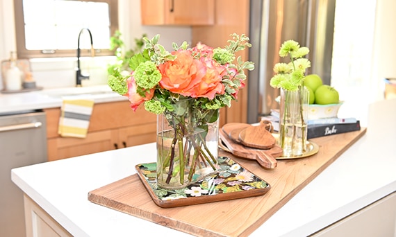 Closeup of kitchen island with white quartz countertop holding butcher block cutting board, glass vase with red flowers and greenery.