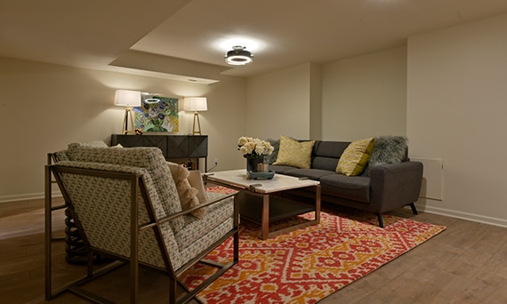 Finished basement with floor tile that looks like wood plank flooring, area rug, brown sofa, stone & metal coffee table, and beige & gray chair.