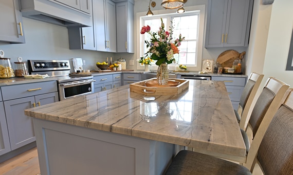 Kitchen with soft blue cabinets, brass pulls, gray quartzite countertop and island, stainless steel farm sink in front of two windows.