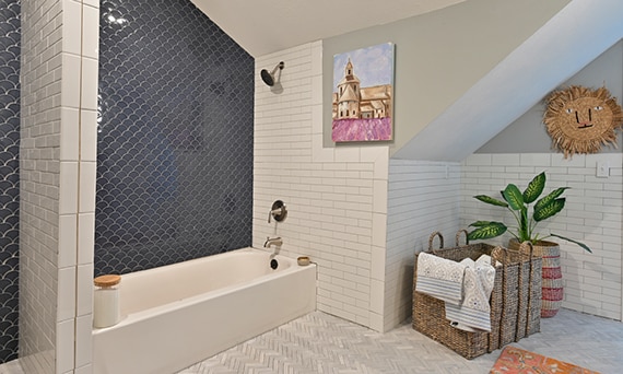 Bathroom with white subway and blue fan-shaped shower tile, gray limestone herringbone floor tile, and wicker laundry basket.