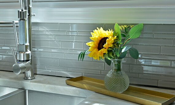 Closeup of kitchen backsplash with gray glass mosaic tile and a glass vase holding a single sunflower on a gray natural quartzite countertop.