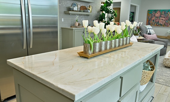 Closeup of white tulips on a kitchen island of taupe natural quartzite countertop with floating shelves, and taupe glass mosaic backsplash in the background.