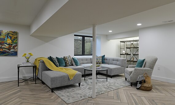 Basement sitting area with herringbone wood look tile flooring, gray sectional sofa, gray chair, coffee table, and shelving.