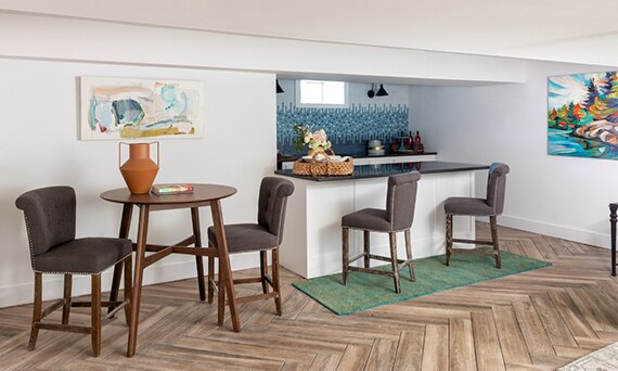 Basement with herringbone wood look tile flooring, table & chairs, wet bar with mosaic backsplash & black quartz countertop.
