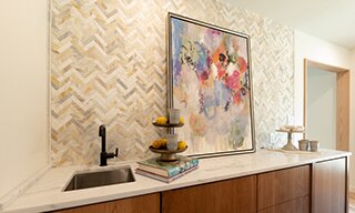 Dining room with beige & tan herringbone marble mosaic wall tile, white & gray veining countertop over wood cabinets, and abstract painting of flowers.