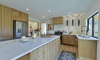 Kitchen island with white marble slab countertop, natural wood cabinets, stainless steel refrigerator and gas oven & stove top with wood vent hood.