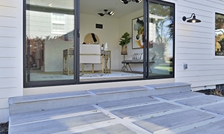 Outdoor patio with gray stone look 2CM porcelain pavers set in white gravel, accordion glass doors with a black frame leading to an indoor sunroom.
