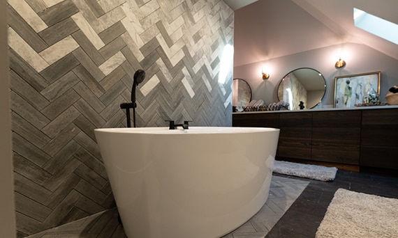Freestanding soaking tub in front of chevron backsplash of white & gray marble tile blended with gray limestone tile, and matte black faucet.