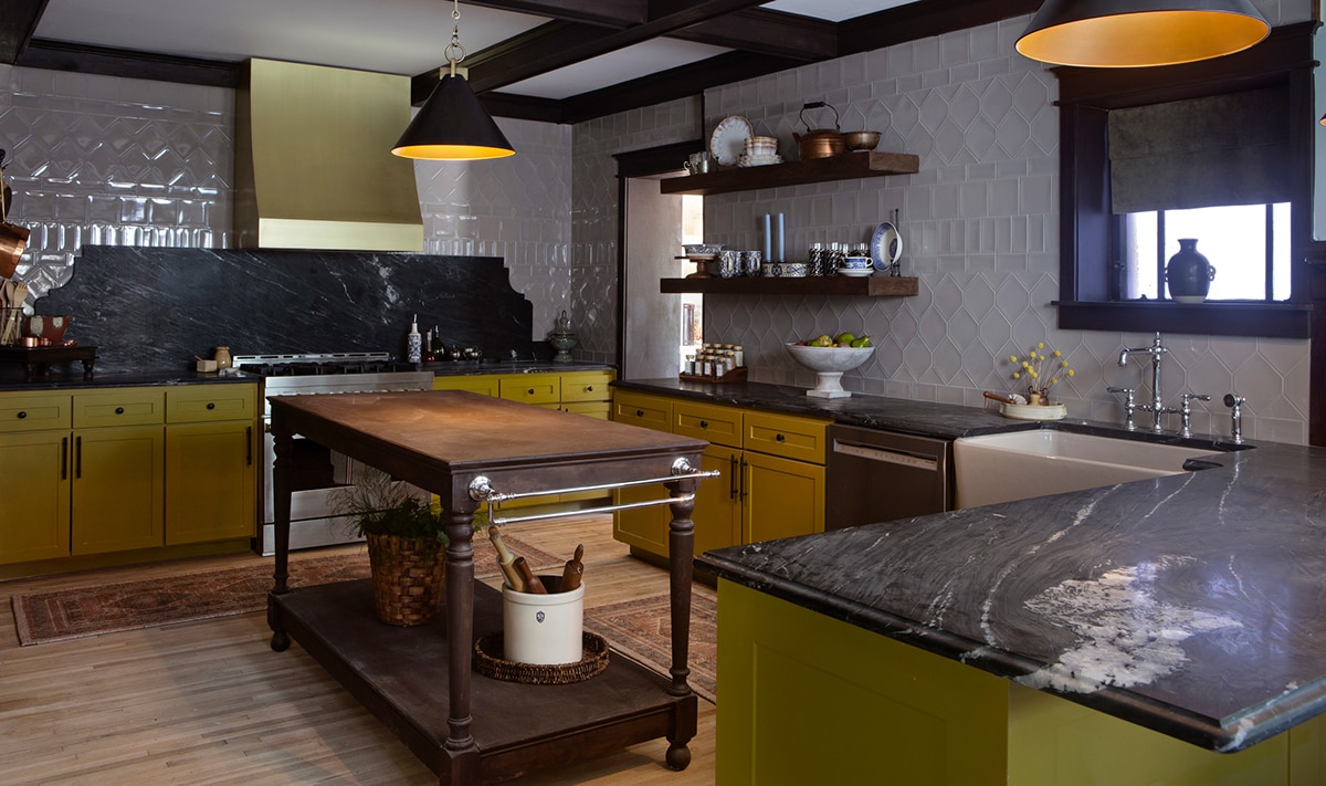 Kitchen with yellow cabinets, light wood floors, tan tile in various sizes on the walls, and a black marble look slab countertop and backsplash.