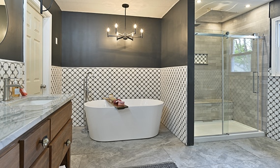 Bathroom with free standing bathtub in front of wainscoting of white marble tile with gray rings and shower with gray tile and niche with matching marble tile with rings.