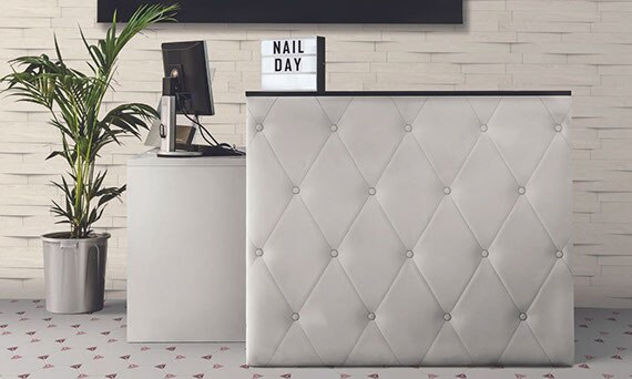 Nail salon with wall with tile that looks like white stacked stone, white tufted leather check-in desk, white, maroon & pink hexagon floor tile.