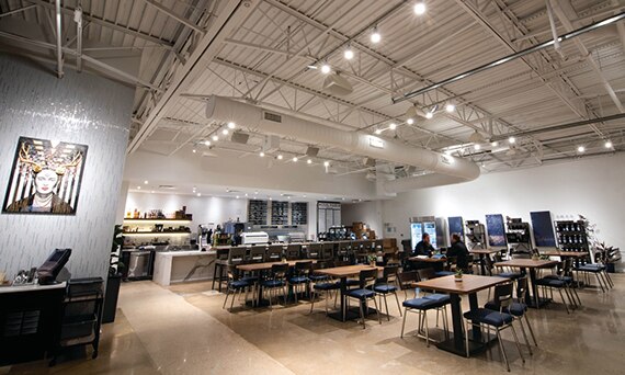 Coffee shop with white industrial style ceiling of exposed air conditioning vent and track lighting, tan concrete flooring, and table with black chairs.
