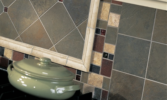 Closeup of kitchen backsplash of green, teal, rust, and beige tile that looks like slate stone and green ceramic pot on stovetop.