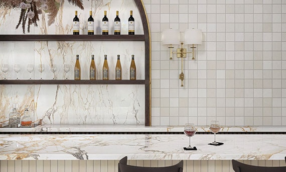 Elegant restaurant bar with porcelain slab countertop that looks like white and golden-brown marble, beige tile wall tile, shelves holding bottles of wine.