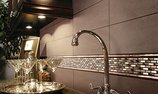 Kitchen backsplash close up with solid taupe four by twelve inch tiles and accent of mixed metals mosaic. Sink faucet and martini glasses in the foreground.