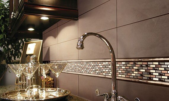 Kitchen backsplash close up with solid taupe four by twelve inch tiles and accent of mixed metals mosaic. Sink faucet and martini glasses in the foreground.