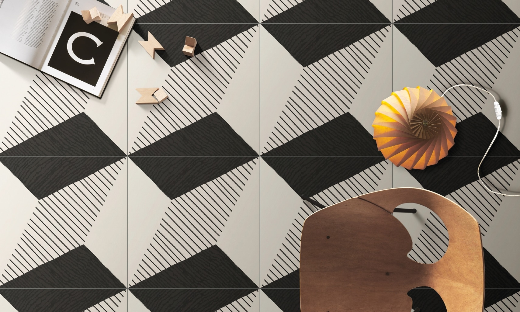 Bird’s eye view of black and white geometric floor tile, small floor lamp next to wooden chair, wood blocks and open book.
