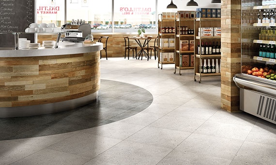 Small market and deli shop with industrial style shelving on the side containing products. Cafe tables in the background. Concrete-look porcelain tile on the floor.