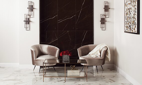 Lounge area with white & gray floor tile and black wall tile that look like marble, beige velvet chairs behind brass and glass coffee table.