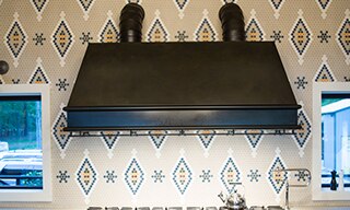 Closeup of kitchen stainless steel gas stovetop & bronze vent hood, light brown quartzite countertop, & island, backsplash of custom tan, white, and tan penny round mosaic tile.