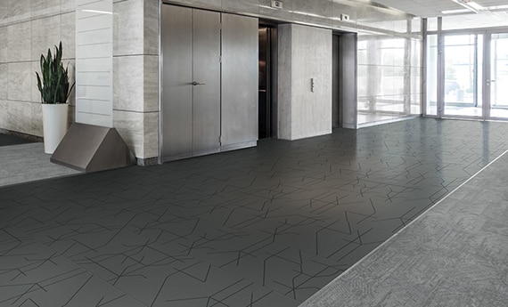 Office building elevator corridor with light and dark gray tile flooring with black design accents, light gray marble walls, and floor to ceiling windows.