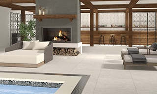 Hotel pool with gray stone-look deck tile, outdoor fireplace with gray tile surround and natural wood mantle, off-white chaise lounge chairs, and outdoor bar.