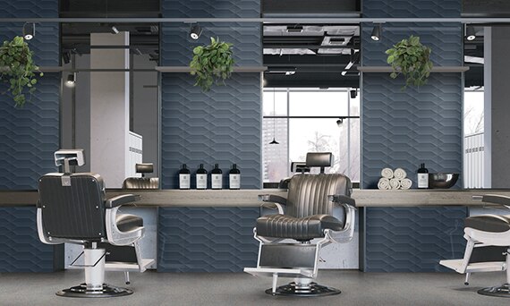 Barber shop with blue textured wall tile, gray terrazzo floor tile, silver leather chairs, large wall mirrors, gray wood countertop holding rolled towels, and hair products.
