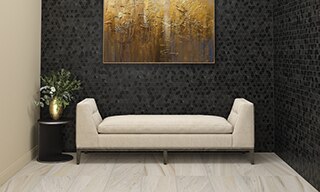 Lobby with beige marble tile flooring, black natural stone mosaic walls, ecru linen chaise lounge, and greenery in a gold vase on a small round side table.