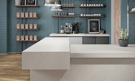 Coffee shop with wood look flooring tiles, off-white stone look quartz countertops, blue diamond-shaped wall tile, and floating shelves holding cups, glasses, and bags of coffee.
