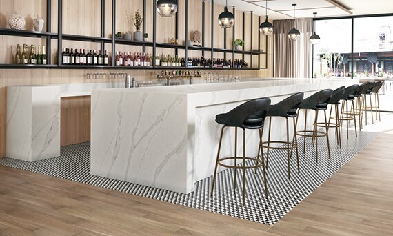 Hotel bar with white & gray marble look quartz countertops, black & white checkered tile mat set in wood look tile flooring, and racks holding wine bottles in front of wood panel wall.