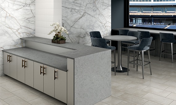 Stadium suite with beige stone look floor tile, gray quartz waterfall countertop, walls covered with porcelain slab that looks like white marble with gray & gold veining.
