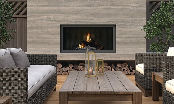 Outdoor patio fireplace with beige porcelain slab surround that looks like travertine, wicker sofa & chair with gray cushions, and wooden coffee table.