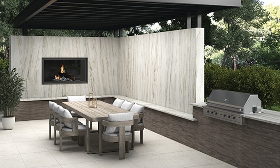 Outdoor patio with pergola, walls with natural quartzite slab, fireplace, wooden table with chairs, grill, and white stone look pavers.