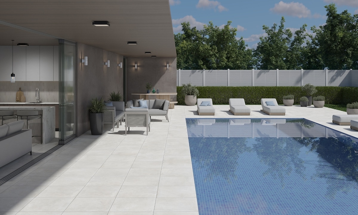Modern home backyard with white marble look paver pool deck, pool with blue mosaic tile, gray seating area under patio cover, and cushioned lounge chairs.