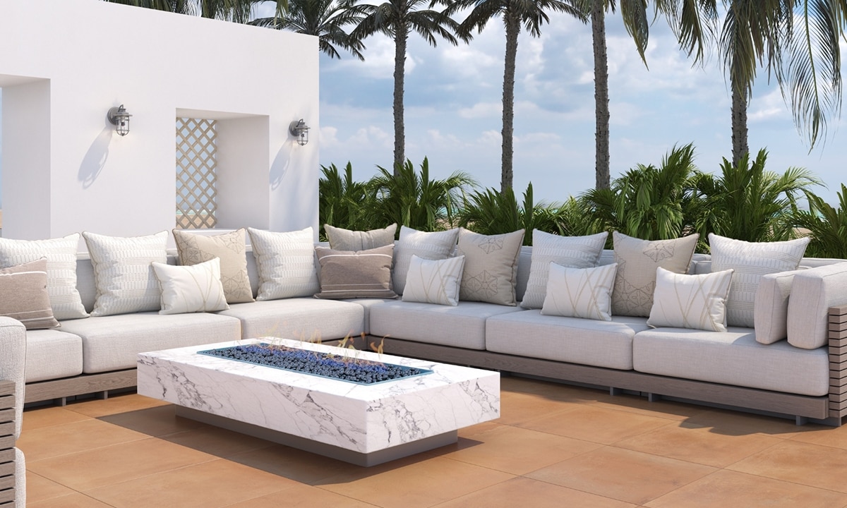 Tropical resort lounge area with terracotta stone look paver flooring, palm trees behind a white sectional sofa that is surrounding a white marble look porcelain firepit.