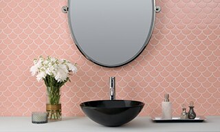 Bathroom vanity with pink fan mosaic backsplash tile, oval mirror, black vessel sink, gray quartz countertop, and bouquet of white carnations.