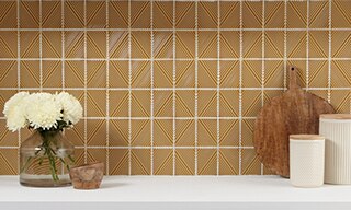 Closeup of kitchen backsplash of burnt orange geometric mosaic tile, white countertop, bouquet of white carnations, and wooden cutting board.