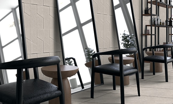 Hair salon with taupe floor tile and wall tile that look like stone, black leather chairs in front of full-length mirrors, and shelving holding hair care products.