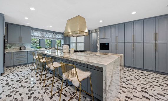 Large kitchen with blue/gray cabinets, white, gray & beige porcelain slab backsplash and waterfall island, and white, gray & beige marble-look mosaic floor tile.