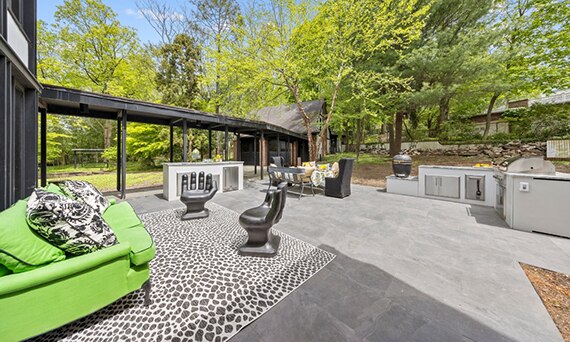 Backyard patio with outdoor kitchen, gray porcelain paver flooring, green couch and two side chairs shaped like hands on black & white animal skin rug.