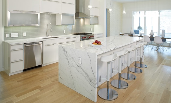Modern kitchen with white & gray waterfall island, white & silver bar stools, wood look floor tiling, white quartz countertops, light green glass backsplash, and white cabinets.